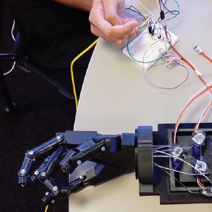 A robotic hand prototype rests on a table, with strings connected to servo motors that control finger movement. A hand is seen bending a flex sensor next to a breadboard, and the robotic hand mimics the bending motion in real-time, indicating successful gesture replication. Wires connect the system components, showcasing a functional flex-sensor-based control setup.